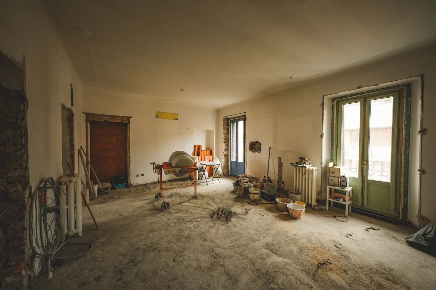 Wohnzimmer im Rohzustand: abgeschlagene Wände, Bauschutt und Werkzeug am Boden, alte Holztür — Originalfoto vor dem virtuellen Home Staging.